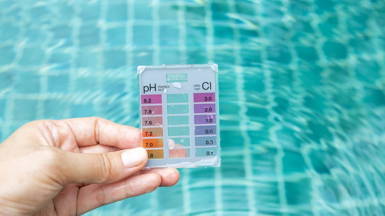 Hand holding a pool water test kit showing pH and chlorine levels above a clear swimming pool.