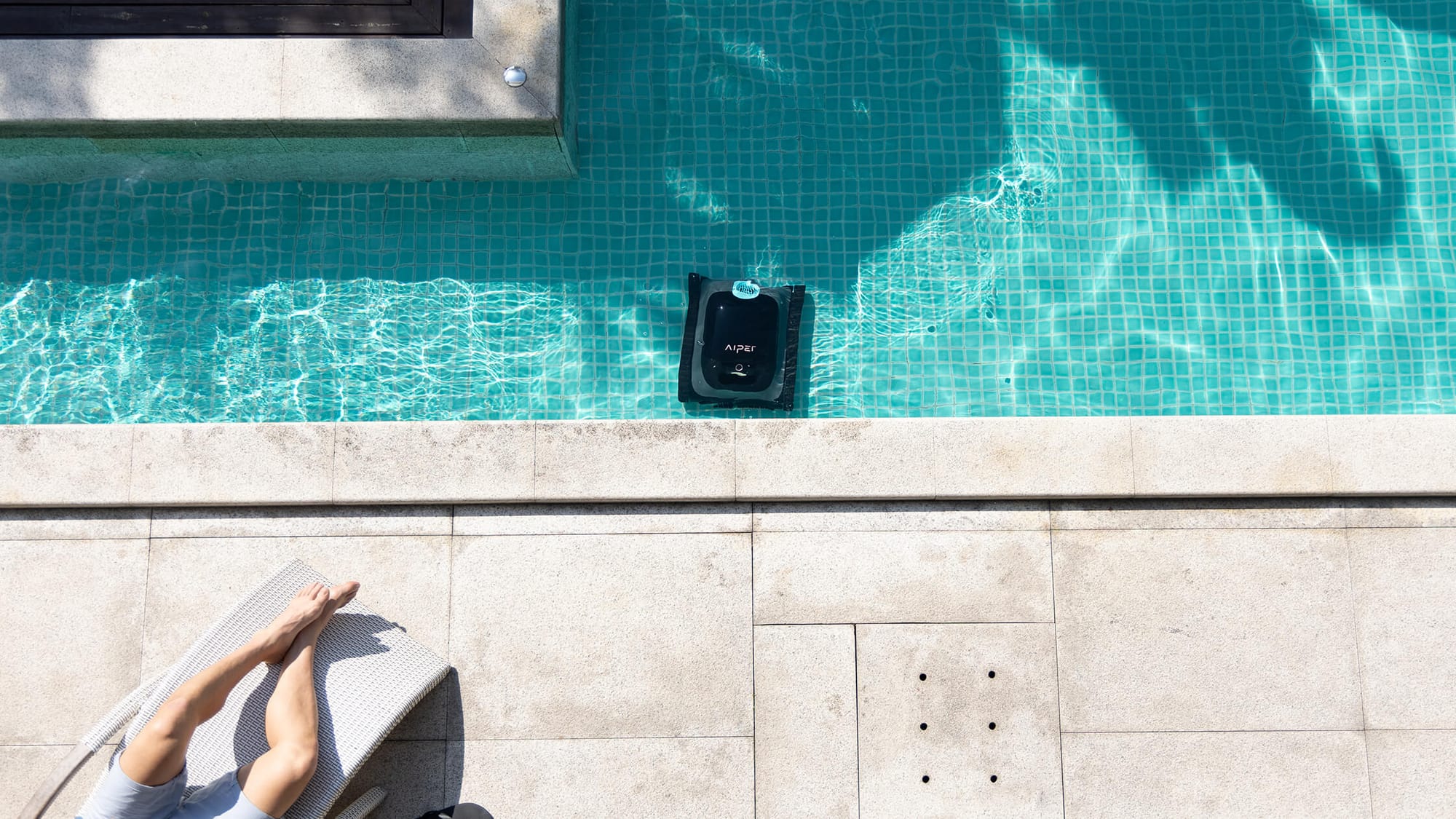 Overhead view of a robotic pool cleaner working on the floor of a pool while a person relaxes poolside.