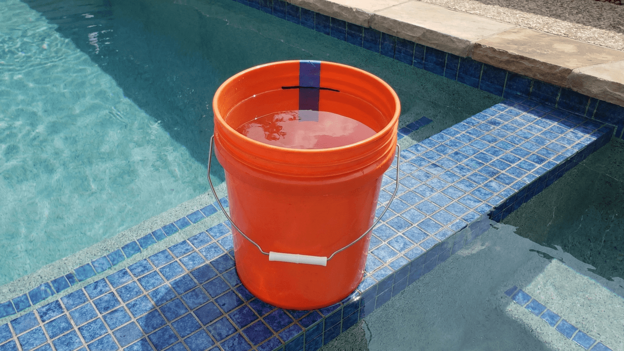 Orange bucket filled with pool water placed on pool step with marked levels for performing the bucket test to detect leaks versus evaporation.